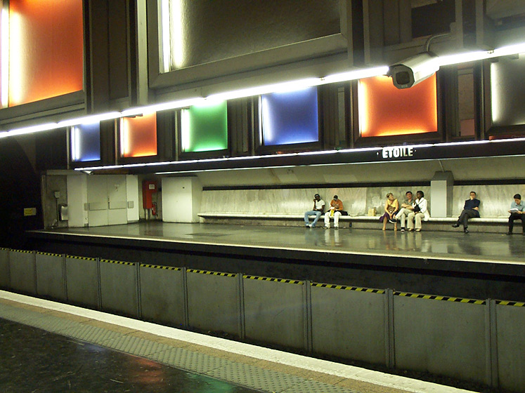 Passengers sit on benches under the brightly lit, colorfully designed Etoile subway station.  Paris, France. 