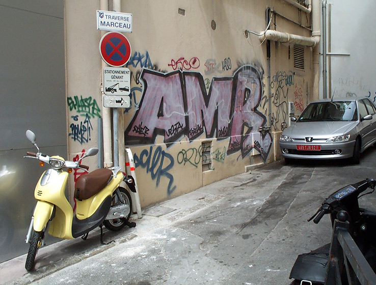 Artistic graffiti adorns the walls of a side street framed by  typically French cars and motorcycles and a tow away warning sign in French. Cannes, South of France.