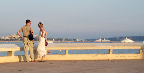 A couple look out over the harbor at the luxury yachts.  The woman flips off her shoe to let some air get to her feet. Cannes, South of France.