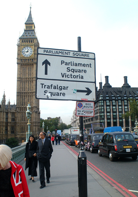 A road sign points the way towards London landmarks near the Houses of Parliament and Big Ben. London taxis and a woman wearing a union jack jacket litter the foreground.    London, U.K..