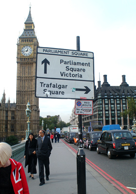 A road sign points the way towards London landmarks near the Houses of Parliament and Big Ben. London taxis and a woman wearing a union jack jacket litter the foreground.  London, U.K..