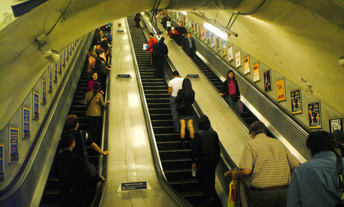 Commuters and tourists ride the escalators past advertising billboards in one of London's tube / subway stations.  London, U.K..