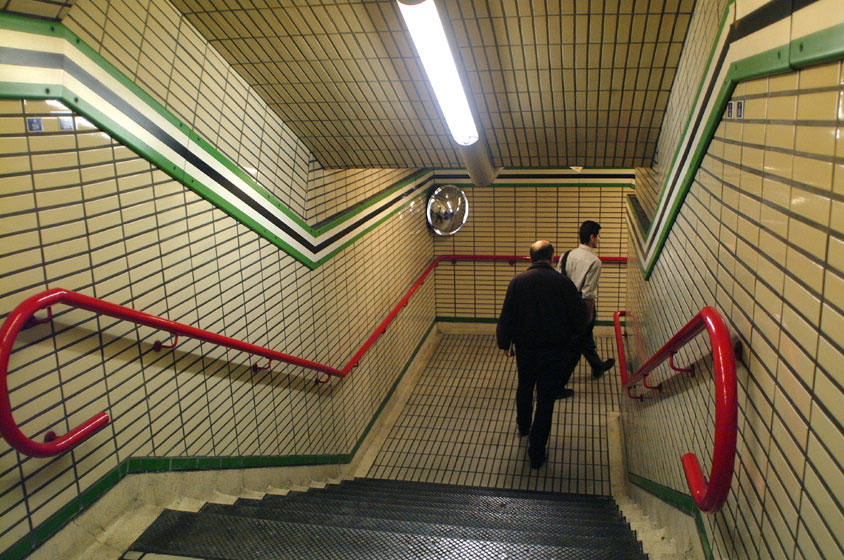 Commuters walk down the stairs to the tube / subway past the security mirrors and the bright red stair railings.  London Underground, London, U.K..